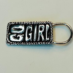 May include: A silver-toned keychain with the word "GIRL" in bold, raised letters. The rectangular pendant has a rope-like border and a small, attached ring. The background is a light, neutral color.