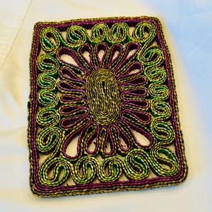 May include: A rectangular woven coaster with a floral design. The coaster features a central oval shape surrounded by purple, green, and gold swirls and petal-like elements. The border is a dark purple.