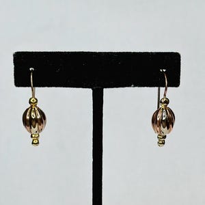 May include: A pair of gold-tone earrings with a drop design. Each earring features a ribbed, rose-gold colored bead suspended from a gold hook. The earrings are displayed on a black stand against a white background.