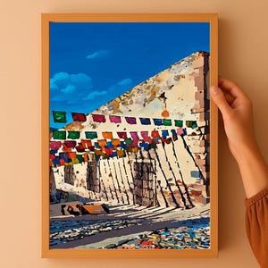 Puede incluir: Una impresión enmarcada de una colorida escena callejera con un cielo azul brillante. La obra de arte representa un edificio con una fachada texturizada, adornada con cuerdas de banderas vibrantes. La impresión es sostenida por una persona.