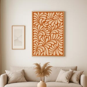Puede incluir: Interior de salón con sofá beige, cojines y mesa de centro de madera. En la pared cuelga una impresión artística abstracta enmarcada, junto a otra obra de arte enmarcada más pequeña. Un jarrón con plantas secas está sobre la mesa. El arte mural tiene un fondo terracota.