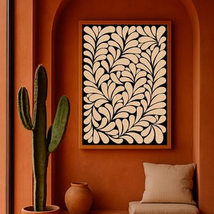 May include: A framed print with a black backdrop and a cream leaf design is displayed on a terracotta wall. A large cactus in a terracotta pot is near a window. A small terracotta vase and a cushion are also present.