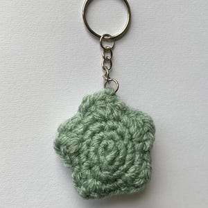 May include: A handmade, sage green crochet keychain in the shape of a five-petal flower. The keychain has a silver-colored metal ring and chain. The flower has a spiral design in the center.