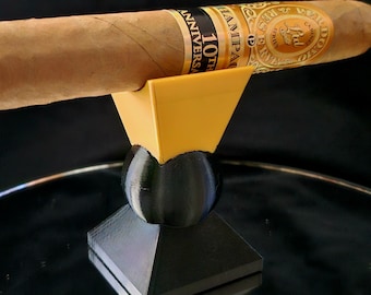May include: A brown cigar rests on a yellow and black cigar stand. The stand has a geometric design with a pyramid base and a spherical midsection. The cigar has a label that reads "10th Anniversary".