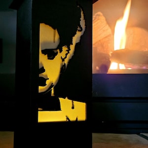 May include: A black metal lantern with a cutout profile of a person's face. The interior glows with a warm yellow light, creating a silhouette effect. The lantern is rectangular and sits on a base.