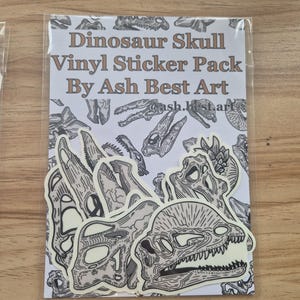 Puede incluir: Un paquete de pegatinas de vinilo con ilustraciones de calaveras de dinosaurios. Las pegatinas tienen varias formas y tamaños, con detallados dibujos lineales en blanco y negro. El embalaje dice "Dinosaur Skull Vinyl Sticker Pack By Ash Best Art."