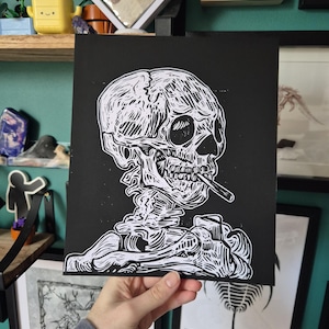 May include: Black and white linocut print of a human skull smoking a cigarette. The skull is detailed with visible bone structure and shading. The artwork is held by a person's hand, with the artist's signature in the bottom right corner.