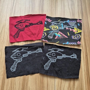 May include: Four fabric patches featuring a black and white illustration of a futuristic ray gun. Two patches are black with white designs, and two are red with black designs. One black patch has a geometric pattern in pink, blue, yellow, and green.