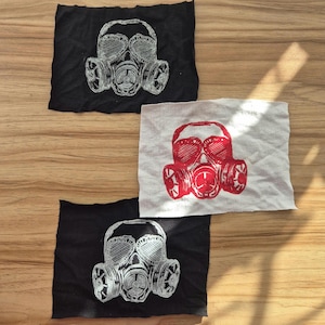 Op de afbeelding: Drie stoffen vierkanten met gasmaskerontwerpen. Twee zwarte vierkanten hebben een witte gasmaskerprint, terwijl een wit vierkant een rood gasmasker toont. Het gasmaskerontwerp omvat een bril en een ademhalingsapparaat.