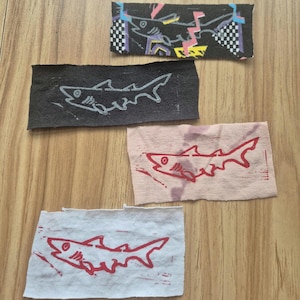 Peut inclure: Quatre carrés de tissu avec des fonds de couleurs différentes et une silhouette de requin imprimée sur chacun. Les requins sont imprimés à l'encre noire, blanche et rouge.