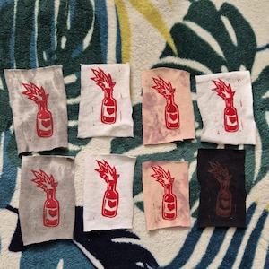 May include: Eight fabric patches featuring a red silhouette of a Molotov cocktail with a heart on the side and flames coming out of the top. The patches are displayed on a patterned surface.