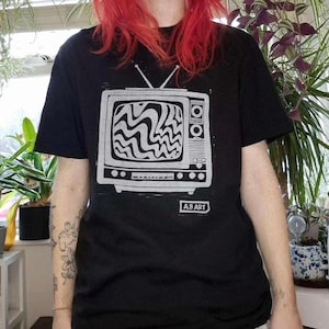 May include: Black t-shirt featuring a white graphic of a vintage television with a wavy, abstract pattern on the screen. The text "A.B. ART" is printed below the graphic.