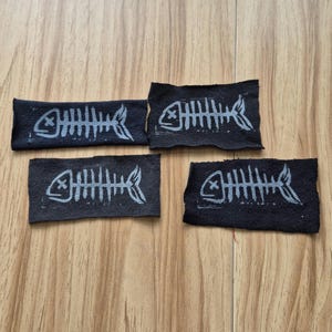May include: Four black fabric patches featuring a white skeleton fish design. Each patch has a white 'X' over the fish's eyes.