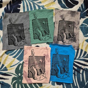 Può includere: Cinque quadrati di tessuto con una stampa in bianco e nero di una chitarra elettrica e un amplificatore. I quadrati sono di diversi colori: grigio, verde, rosa, blu e grigio.