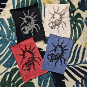 May include: Four fabric patches featuring a black scorpion design. The scorpion has a spiky crown and is printed in black ink on different coloured fabrics: black, white, red, and blue.