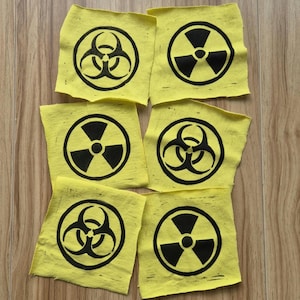 Puede incluir: Seis cuadrados de tela amarilla con símbolos negros de riesgo biológico y radiación. Los símbolos están centrados en los cuadrados, dispuestos sobre una superficie de madera. El símbolo de riesgo biológico es un trébol negro dentro de un círculo.