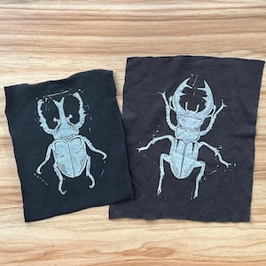 Może przedstawiać: Dwa czarne kwadraty materiału z białymi nadrukami owadów. Owady mają szczegółowe ciała i charakterystyczne rogi. Materiał ma lekko pomarszczoną fakturę, a nadruki wydają się być ręcznie robione.