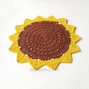 Op de afbeelding: Een handgemaakte, gehaakte zonnebloem doily. Het midden is een diepbruine kleur, omgeven door een ring van openwerk, en omlijst door heldergele, puntige bloemblaadjes. De doily is cirkelvormig en ligt op een wit oppervlak. De tekst "@giftseshop.com" is zichtbaar.
