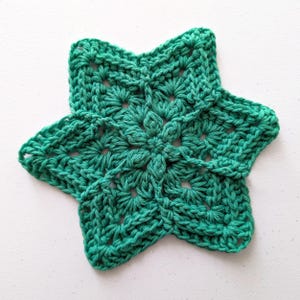 Puede incluir: Estrella de ganchillo verde esmeralda hecha a mano. La estrella tiene seis puntas y un diseño calado detallado. El hilo parece ser un algodón suave, y las puntadas son apretadas y uniformes. La estrella es probablemente un artículo decorativo o parte de un proyecto de artesanía más grande.