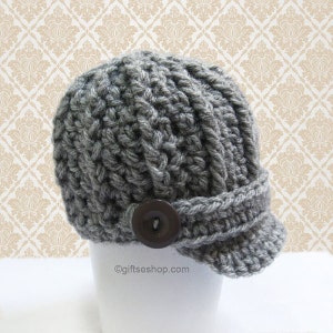 May include: Gray crocheted beanie with a button on the front and a brim. The beanie is on a white surface.