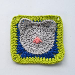Puede incluir: Un cuadrado de ganchillo con un diseño de cara de gato. La cara del gato es gris con una nariz rosa, sobre un fondo azul y verde. Los bordes están bordeados con un hilo verde brillante.