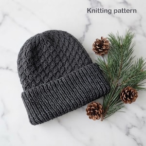 May include: A dark grey knit beanie with a textured diamond pattern and a wide ribbed cuff. The text "Knitting pattern" is visible in the upper right corner, accompanied by a sprig of green pine needles and three brown pinecones.