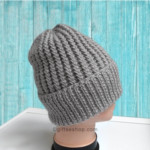 May include: Gray knitted beanie hat with a ribbed texture. The hat is on a mannequin head against a blue wooden background.