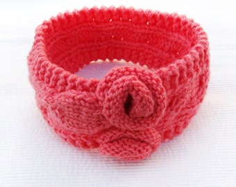 Könnte beinhalten: Ein rosa gestricktes Stirnband mit einem großen Blumenakzent. Das Stirnband ist aus weichem, dehnbarem Garn gefertigt und perfekt, um die Haare aus dem Gesicht zu halten.