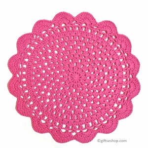 Könnte beinhalten: Eine rosa gehäkelte Deckchen mit einem gewellten Rand. Die Deckchen ist mit einem zarten Stichmuster gearbeitet und hat eine runde Form.