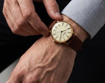 Könnte beinhalten: Eine goldfarbene Armbanduhr mit einem runden cremefarbenen Zifferblatt, schwarzen arabischen Ziffern und dünnen schwarzen Zeigern. Die Uhr verfügt über ein braunes Lederarmband mit sichtbaren Nähten und einer Metallschließe am Handgelenk.