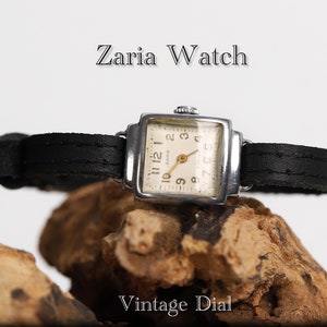 May include: A silver square-shaped wristwatch with a black leather band. The watch face is white with black Roman numeral hour markers and a small second hand. The watch face also has the text "ZAPCH" in black. The watch is sitting on a piece of wood. The text "Zaria Watch" is above the watch and the text "Vintage Dial" is below the watch.