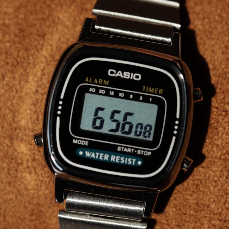 K&ouml;nnte beinhalten: Eine silberfarbene Casio Digitaluhr mit rechteckigem schwarzem Zifferblatt, digitaler Anzeige von 6:56:08, Alarm- und Timer-Funktionen sowie einem Metallgliederarmband. Das Zifferblatt tr&auml;gt die Aufschriften CASIO, ALARM, TIMER und WATER RESIST.