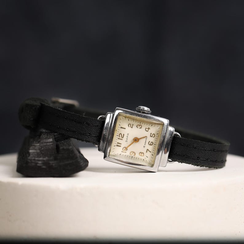May include: A silver square wristwatch with a black leather band. The watch face is white with black Roman numeral hour markers and black Arabic numeral minute markers. The watch has a small second hand.