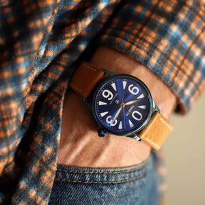 Peut inclure: Une montre-bracelet à cadran bleu avec des chiffres et des aiguilles blancs, et un bracelet en cuir marron. La montre est portée au poignet, avec une chemise à carreaux et un jean en arrière-plan. Le cadran de la montre porte le mot "PAKETA".