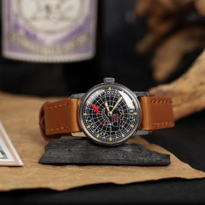 May include: A vintage wristwatch with a black face and a brown leather band. The watch face features a map of the USSR with a red airplane icon.