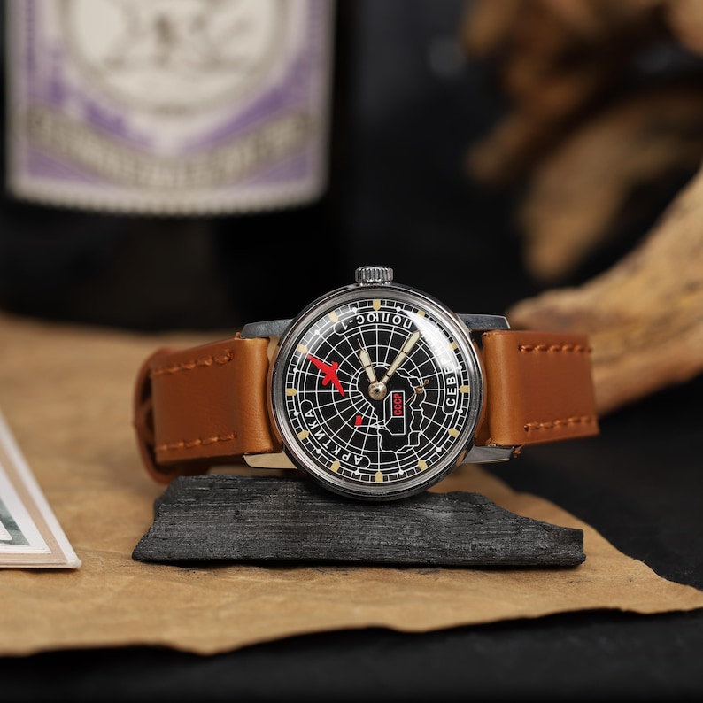 May include: A vintage wristwatch with a black face and a brown leather band. The watch face features a map of the USSR with a red airplane icon.