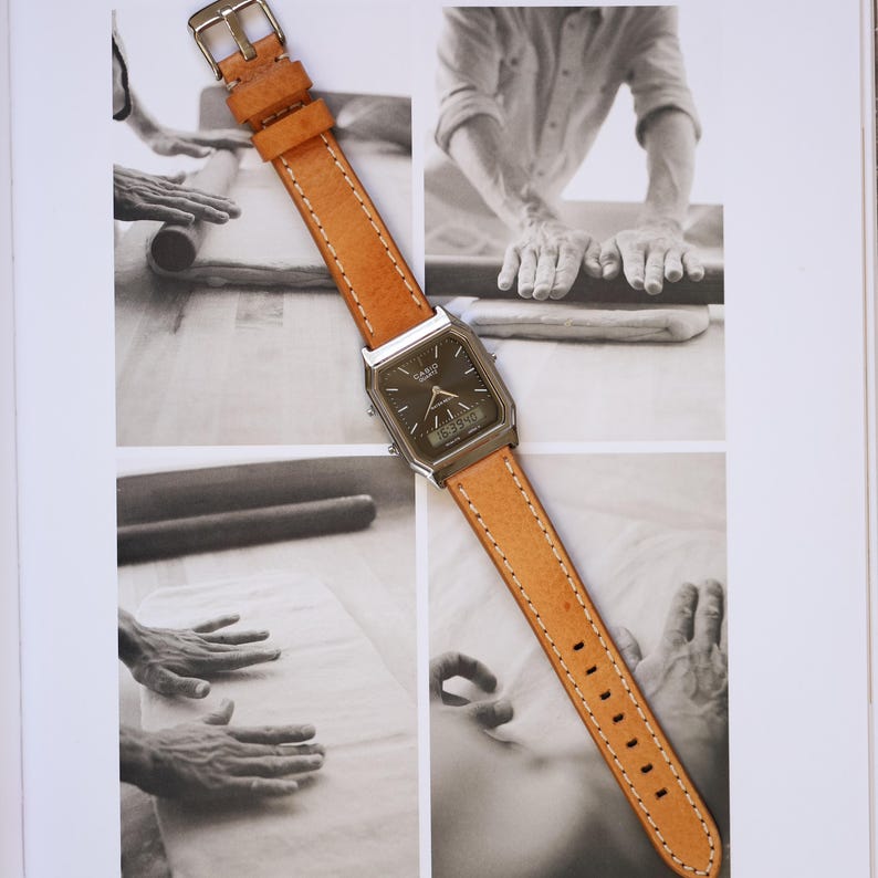 May include: A vintage Casio watch with a brown leather strap. The watch has a silver-tone case and a dark face with both analog hands and a digital display. The light brown leather strap is stitched with white thread and has a silver buckle.