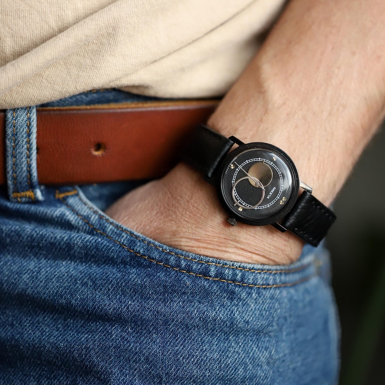 May include: A black wristwatch with a leather band is displayed. The watch face has a unique design with gold accents. The watch is worn on a wrist, with the person wearing blue jeans and a brown leather belt.