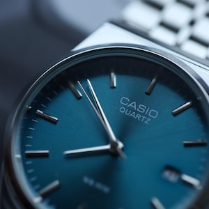 May include: Close-up of a Casio Quartz watch with a teal blue dial and silver hands. The watch has a silver band and a minimalist design. The watch face displays the brand name.