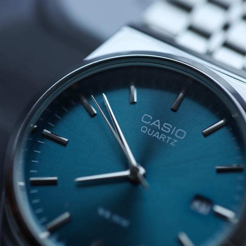 May include: Close-up of a Casio Quartz watch with a teal blue dial and silver hands. The watch has a silver band and a minimalist design. The watch face displays the brand name.