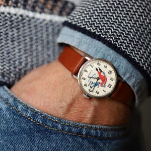 Könnte beinhalten: Eine runde analoge Armbanduhr mit hellbraunem Lederarmband und silberfarbenem Gehäuse. Das cremefarbene Zifferblatt zeigt schwarze arabische Ziffern, dünne schwarze Zeiger und eine grafische Abbildung einer Eislauffigur. Der Text Zarya ist aufgedruckt.
