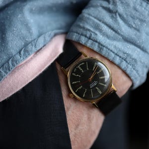 May include: A vintage wristwatch with a black face and gold-colored case. The watch has a black mesh band and is worn on a wrist. The watch face features gold-colored hands and markers. The watch is a classic accessory.