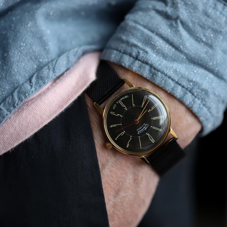 May include: A vintage wristwatch with a black face and gold-colored case. The watch has a black mesh band and is worn on a wrist. The watch face features gold-colored hands and markers. The watch is a classic accessory.