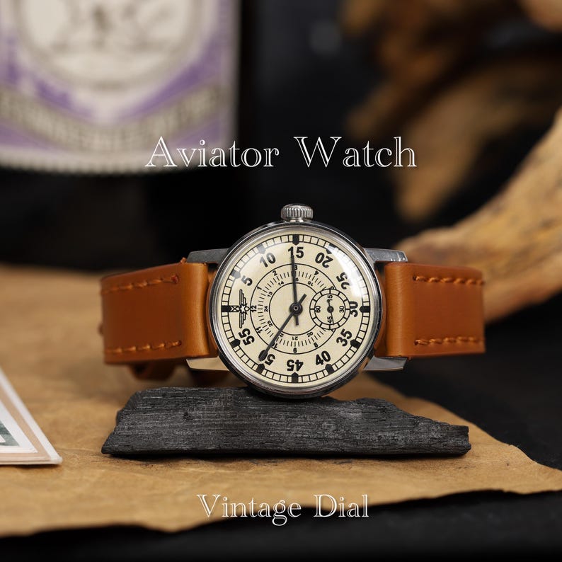 May include: A vintage aviator watch with a white face and black markings. The watch has a brown leather band and a silver case. The watch face has a small subdial at the 3 o'clock position. The watch face also has the text "Aviator Watch" and "Vintage Dial".