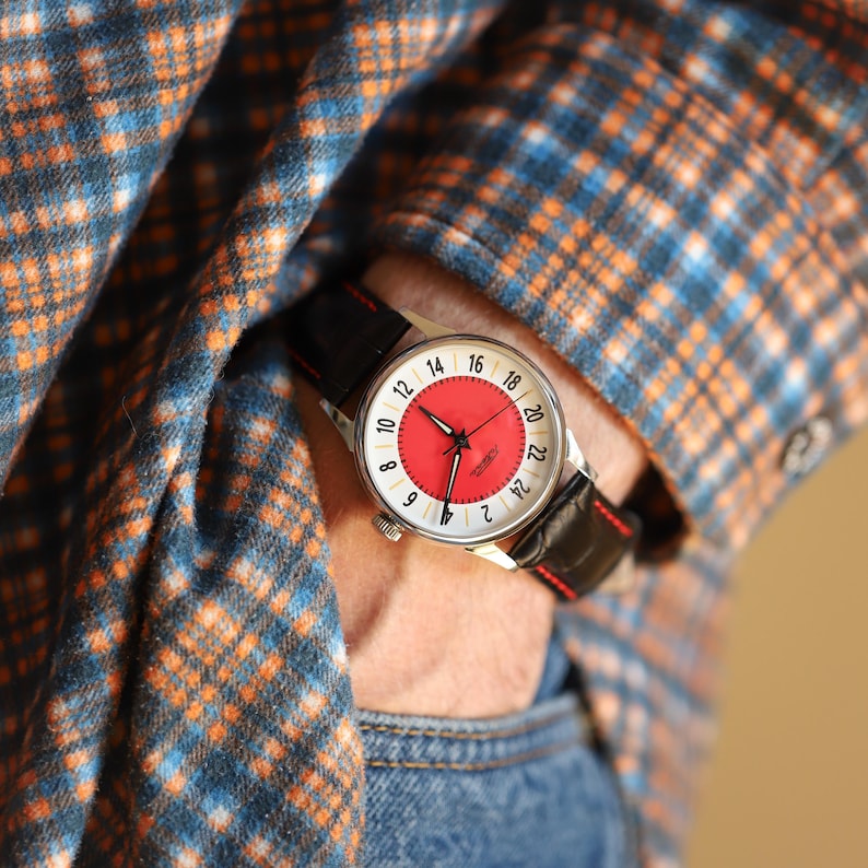 May include: A wristwatch with a red face and black leather band. The watch has a silver case and white numerals. The band has red stitching. The watch is being worn on a wrist, with a plaid shirt in the background.