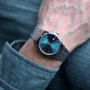 May include: A silver-toned wristwatch with a blue dial. The watch has a rectangular face and a linked metal band. The watch is on a wrist, with a gray long-sleeved shirt in the background. The watch face has the brand name "Casio Quartz".