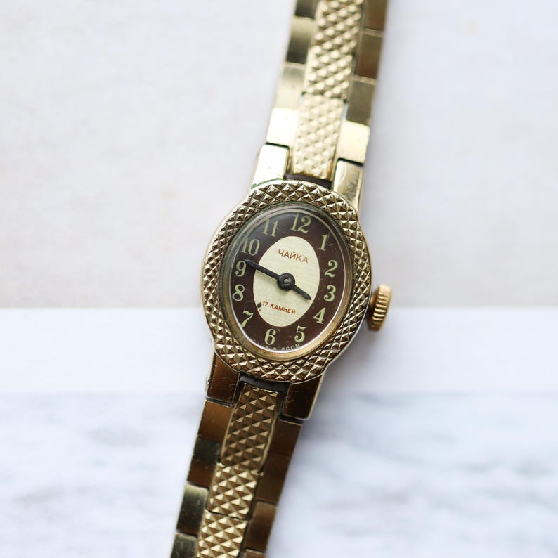 Puede incluir: Un reloj de pulsera vintage dorado con una esfera ovalada. La esfera del reloj es marr&oacute;n con n&uacute;meros y manecillas blancas. La correa del reloj es de color dorado texturizado. La esfera del reloj tiene la palabra "ЧАЙКА" impresa.