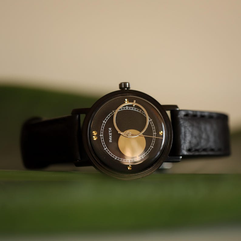 May include: A black wristwatch with a round face and a black leather band. The watch face features gold accents and the word "PAKETA". The watch is set against a blurred green and beige background.