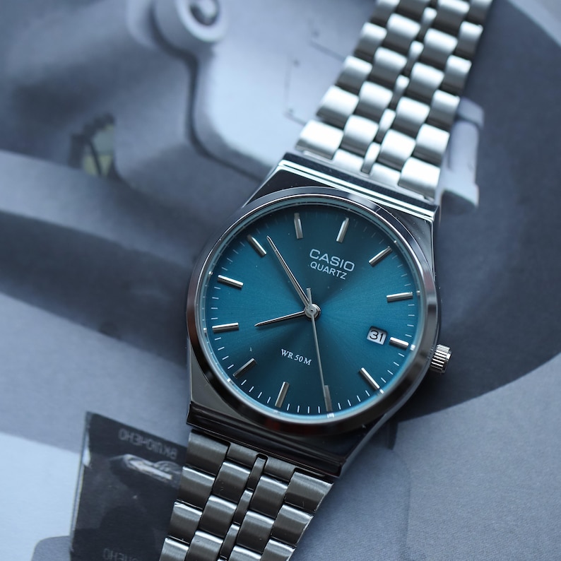 May include: A silver-toned Casio Quartz watch with a teal blue dial and a date display at the 3 o'clock position. The watch has a stainless steel band and the text "WR 50M" is visible on the dial.