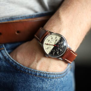 May include: A wristwatch with a silver case and a two-tone dial, split between cream and black. The watch has a brown leather strap. The watch face displays numbers and markings for timekeeping.